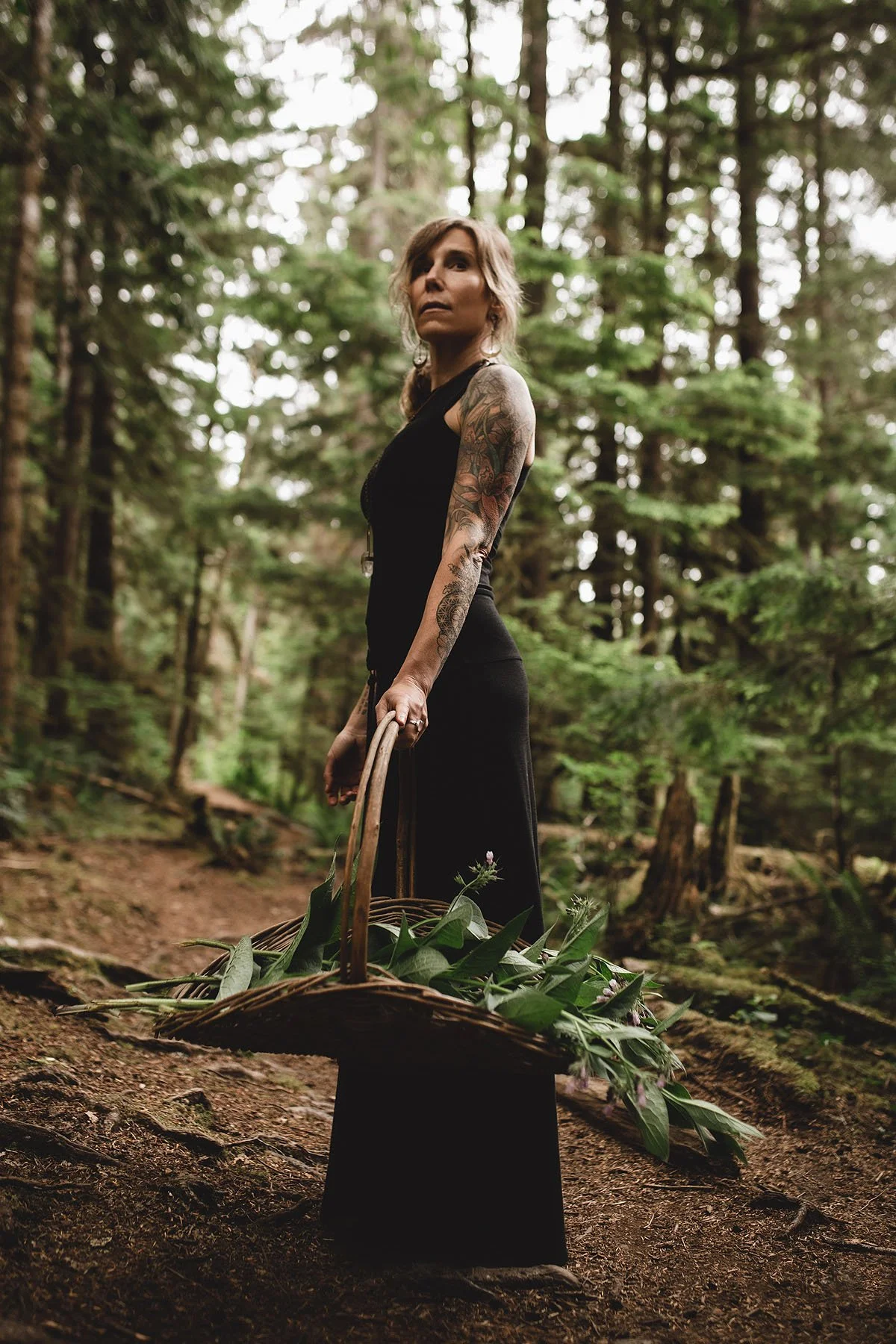 Natalie Rousseau stands in a forest holding a basket filled with gathered green leaves, surrounded by tall trees and woodland ground cover.