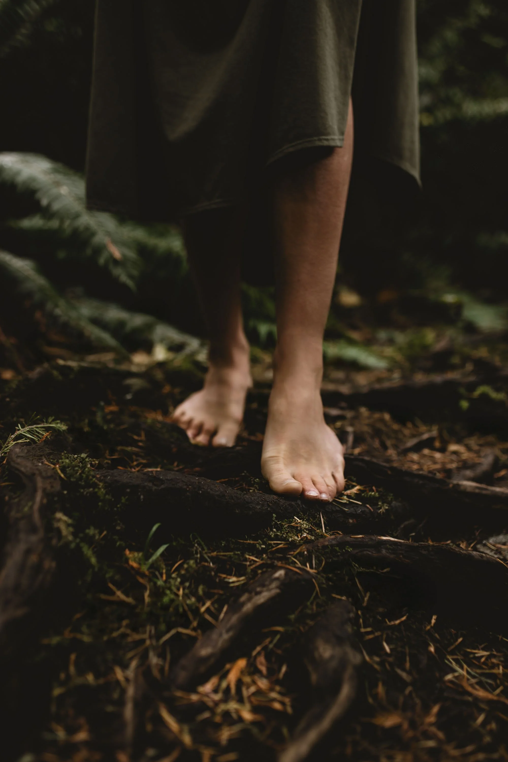 Bare feet walking on a forest floor covered with roots and moss, with a person wearing a loose dark shirt or robe.