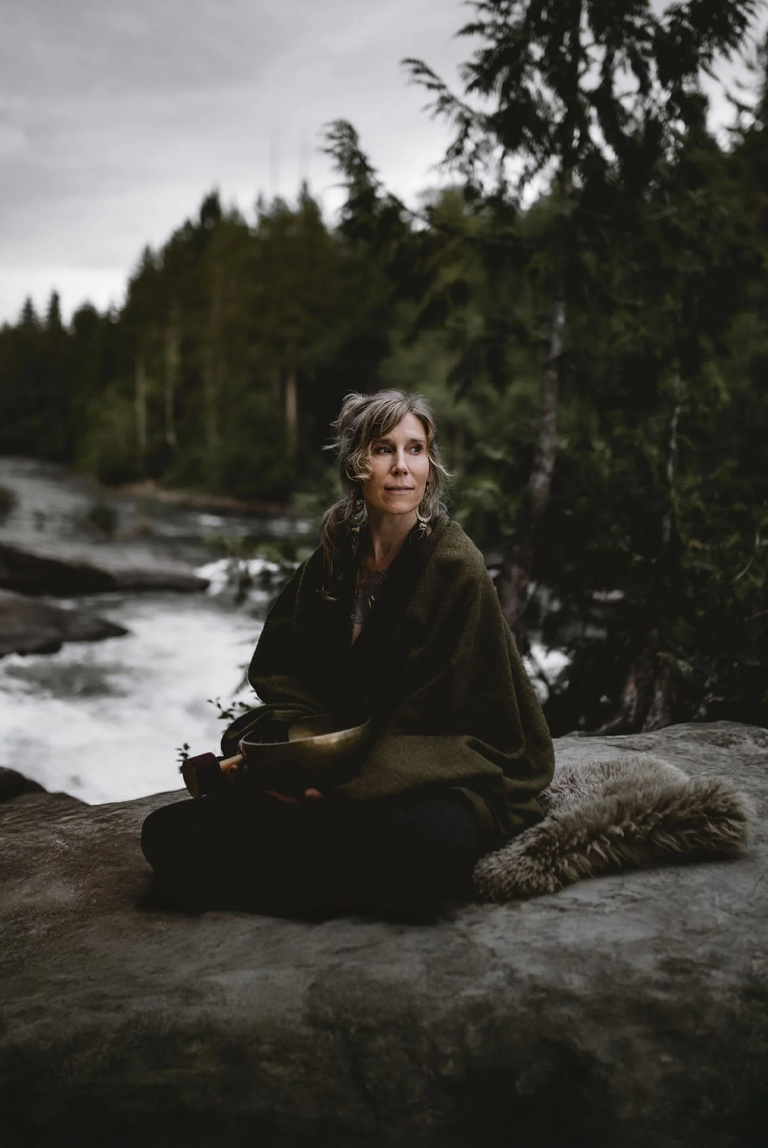 Natalie Rousseau sits wrapped in a shawl on a rock beside a flowing river in a forest, holding a metal bowl and looking off into the distance.