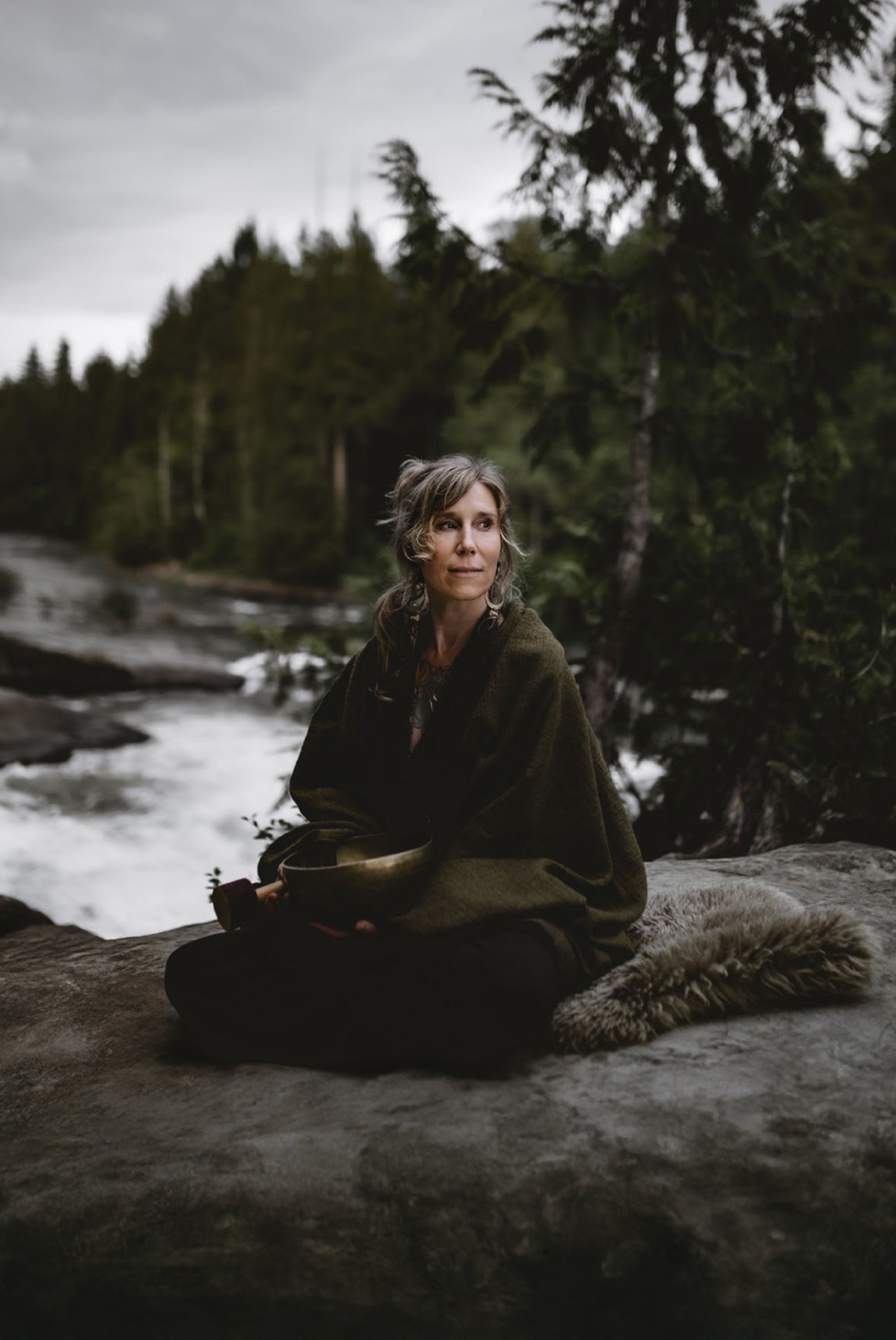 Natalie Rousseau sits wrapped in a shawl on a rock beside a flowing river in a forest, holding a metal bowl and looking off into the distance.