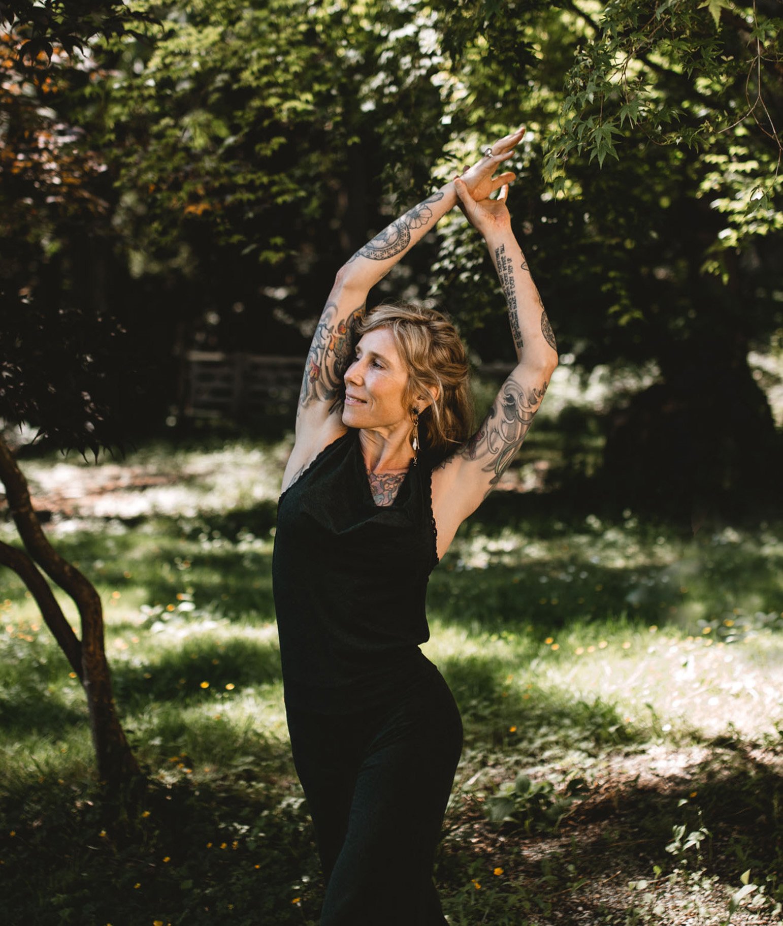 Natalie Rousseau practicing a gentle yoga posture outdoors with arms raised among trees, expressing somatic awareness and grounded movement