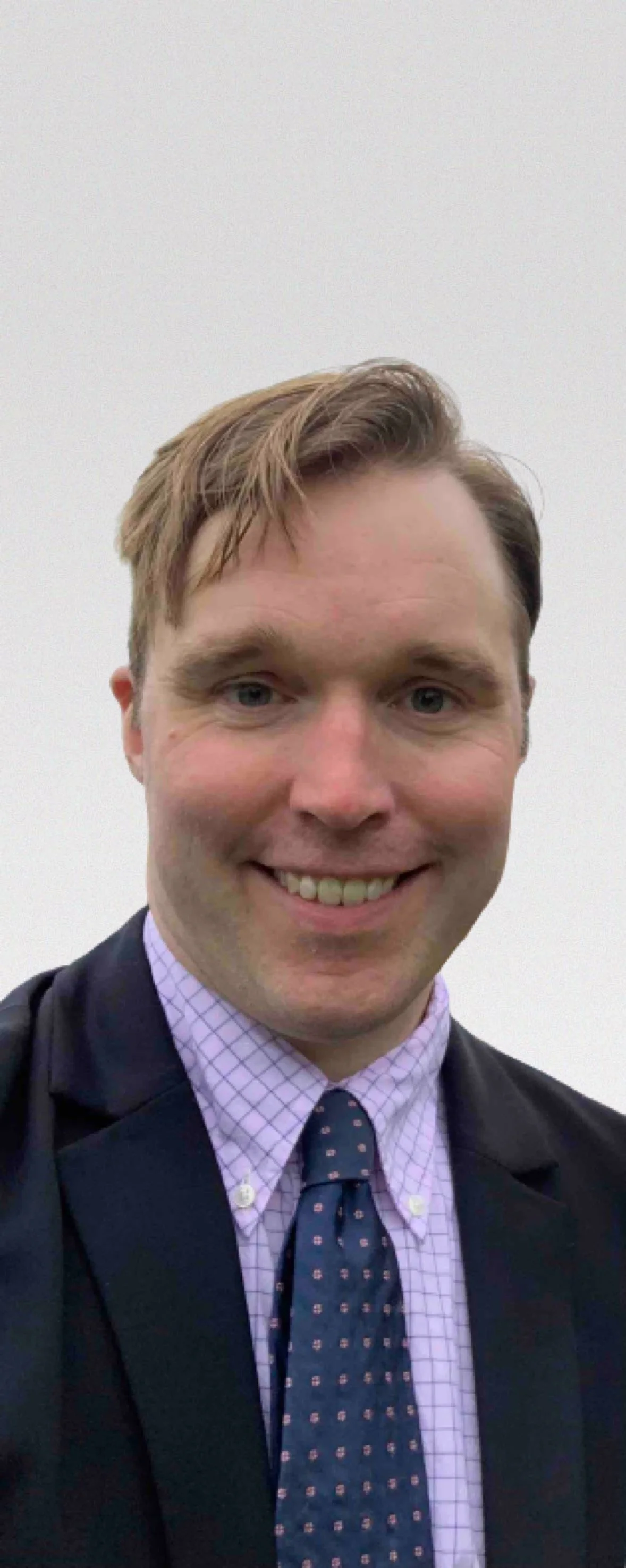 A young man with light brown hair is smiling in a professional setting, dressed in a suit and tie, with a white grid shirt and a patterned dark blue tie, against a plain white background.