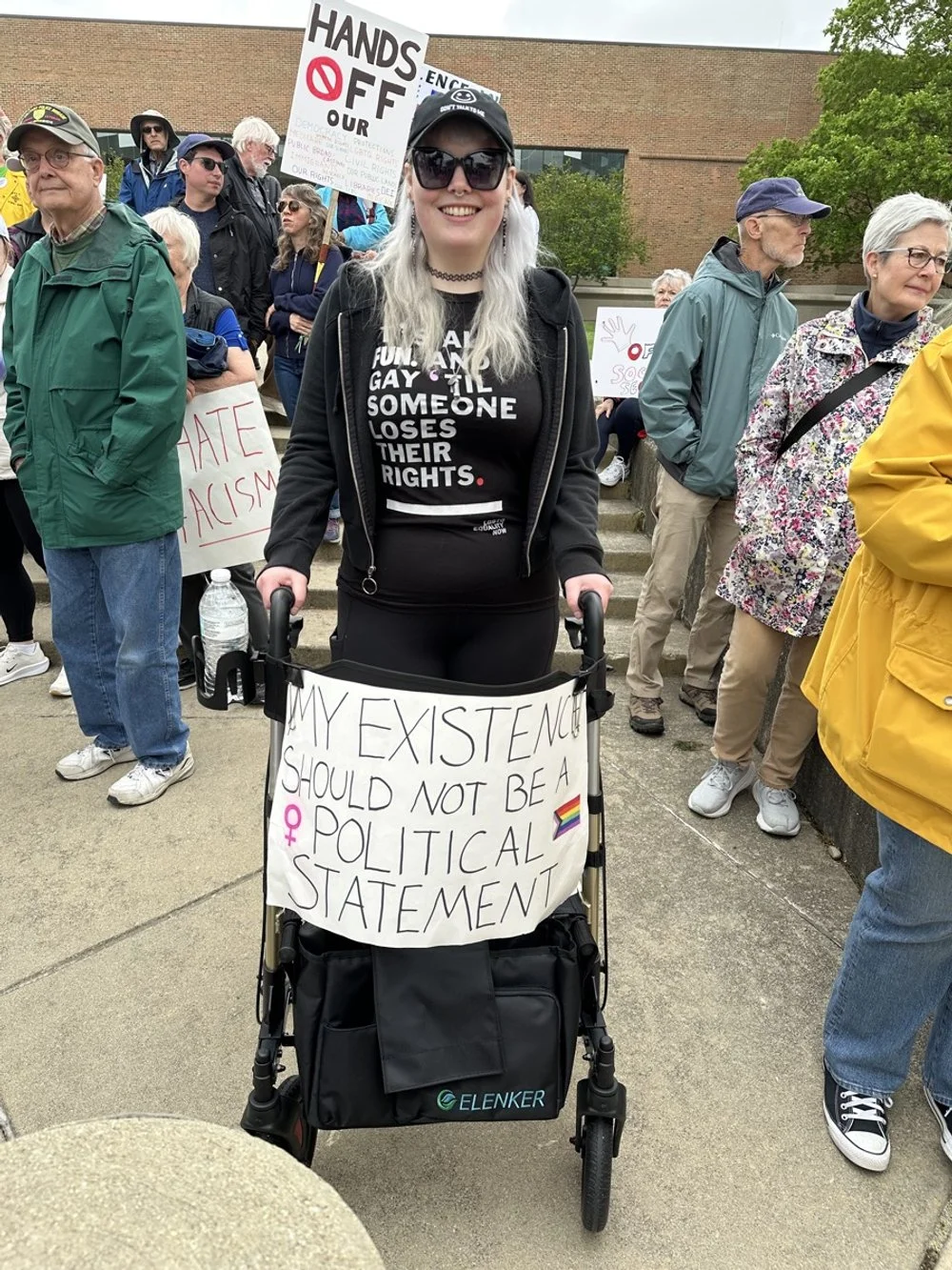 A woman in a black hoodie with white text, wearing sunglasses and having white hair, standing with a walker that has a sign reading 'My existence should not be a political statement' with a rainbow flag and female gender symbol. She is part of a crow