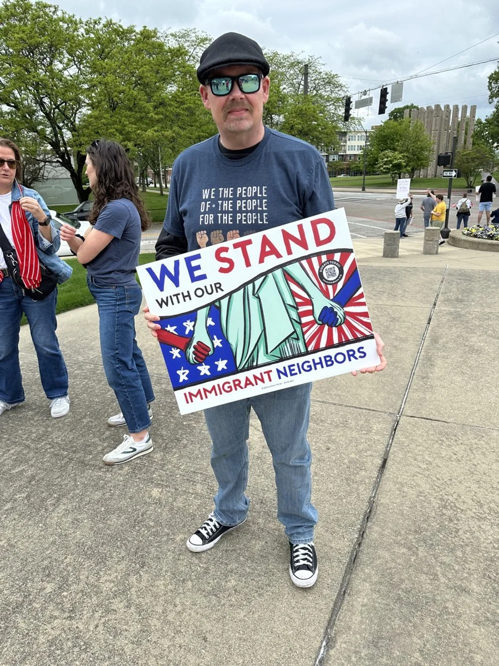 Man wearing sunglasses, a black hat, and a blue T-shirt standing outdoors during a protest or rally, holding a colorful sign that reads "We Stand with Our Immigrant Neighbors" with illustrations of diverse people. Several other people are visible in