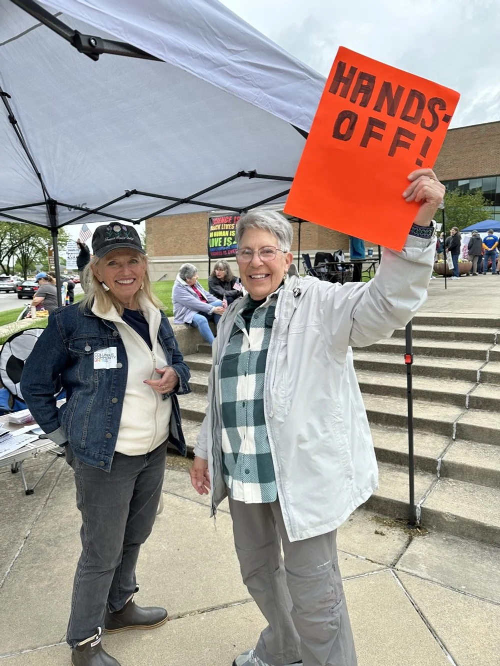 Two women smiling at an outdoor protest or rally under a tent; one holding an orange sign that says "HANDS-OFF!"