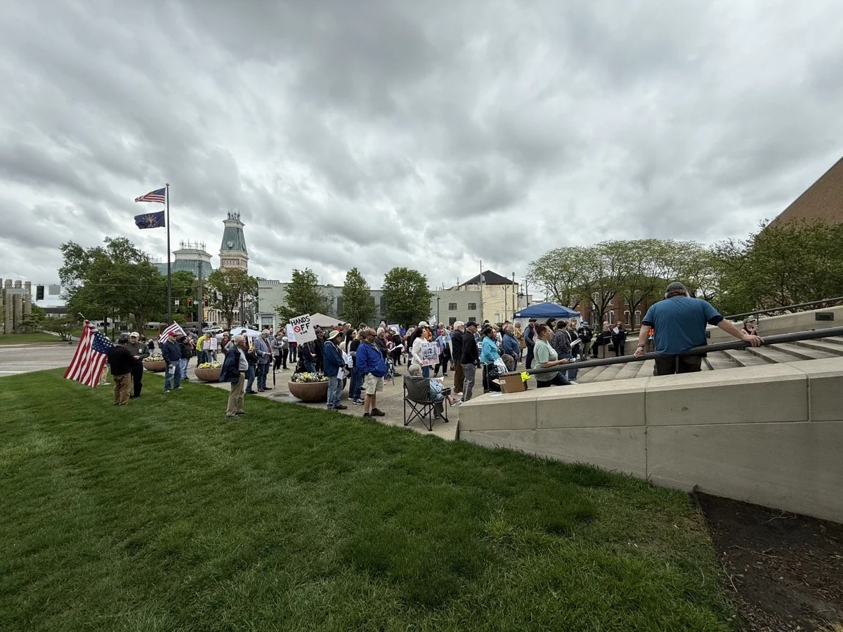 A group of people gathered outdoors, some holding American flags and signs, in front of a historic-looking building with a clock tower under a cloudy sky.