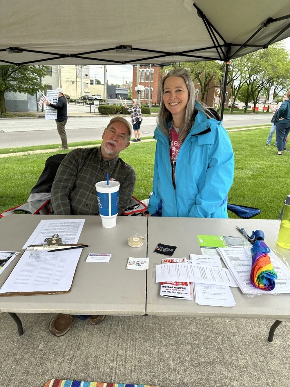 Two smiling women at a booth outdoors under a canopy, with various papers, pamphlets, and rainbow-colored items on the table, and other people holding signs in the background.