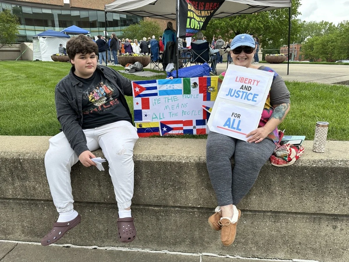 Two people sitting on a concrete ledge at a protest or rally. The boy on the left is holding a small piece of paper. The woman on the right is smiling and holding a sign that reads 'Liberty and Justice for All.' Between them, there is a colorful bann