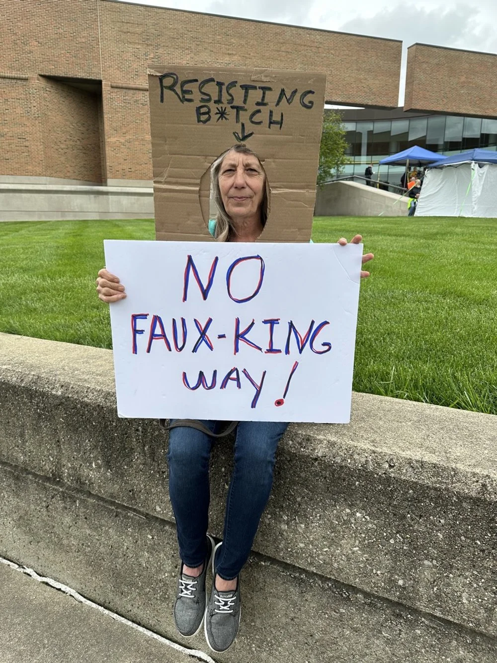 A woman sitting on a concrete ledge holds a white protest sign that says 'NO FAUX-KING WAY!' and a cardboard cutout framing her face with handwritten words 'RESISTING B*TCH' above. She appears at an outdoor protest or rally.