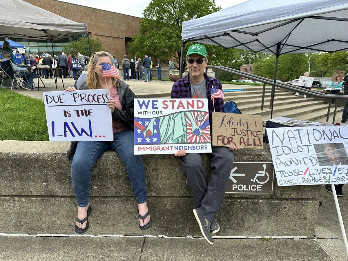 Two protesters sitting on a ledge holding signs and small American flags at a rally. The woman on the left holds a sign reading "DUE PROCESS IS THE LAW..." The man on the right holds a sign that says "WE STAND WITH OUR IMMIGRANT NEIGHBORS" and anothe