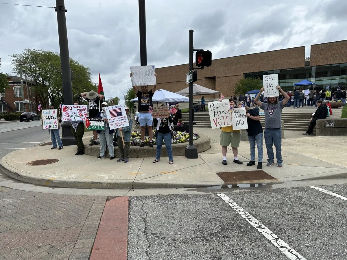 Group of protesters standing on a sidewalk holding signs with messages about social and political issues, in front of a building with stairs, against an overcast sky.
