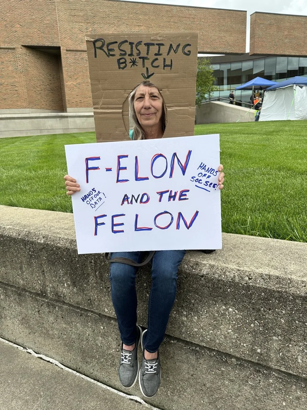 A woman sitting on a concrete ledge holding a protest sign saying 'FELON AND THE FELON' with additional smaller text reading 'HANDS OFF 4 OUR DATA' and 'HANDS OFF 50C SEC'. Behind her, there is a cardboard cutout with the words 'RESISTING B*TCH' and