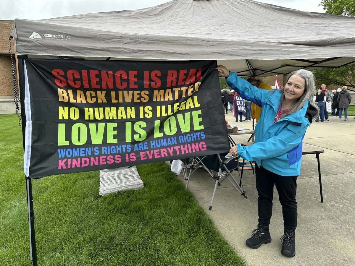 A smiling woman in a blue jacket standing next to a banner with social justice messages at an outdoor event.