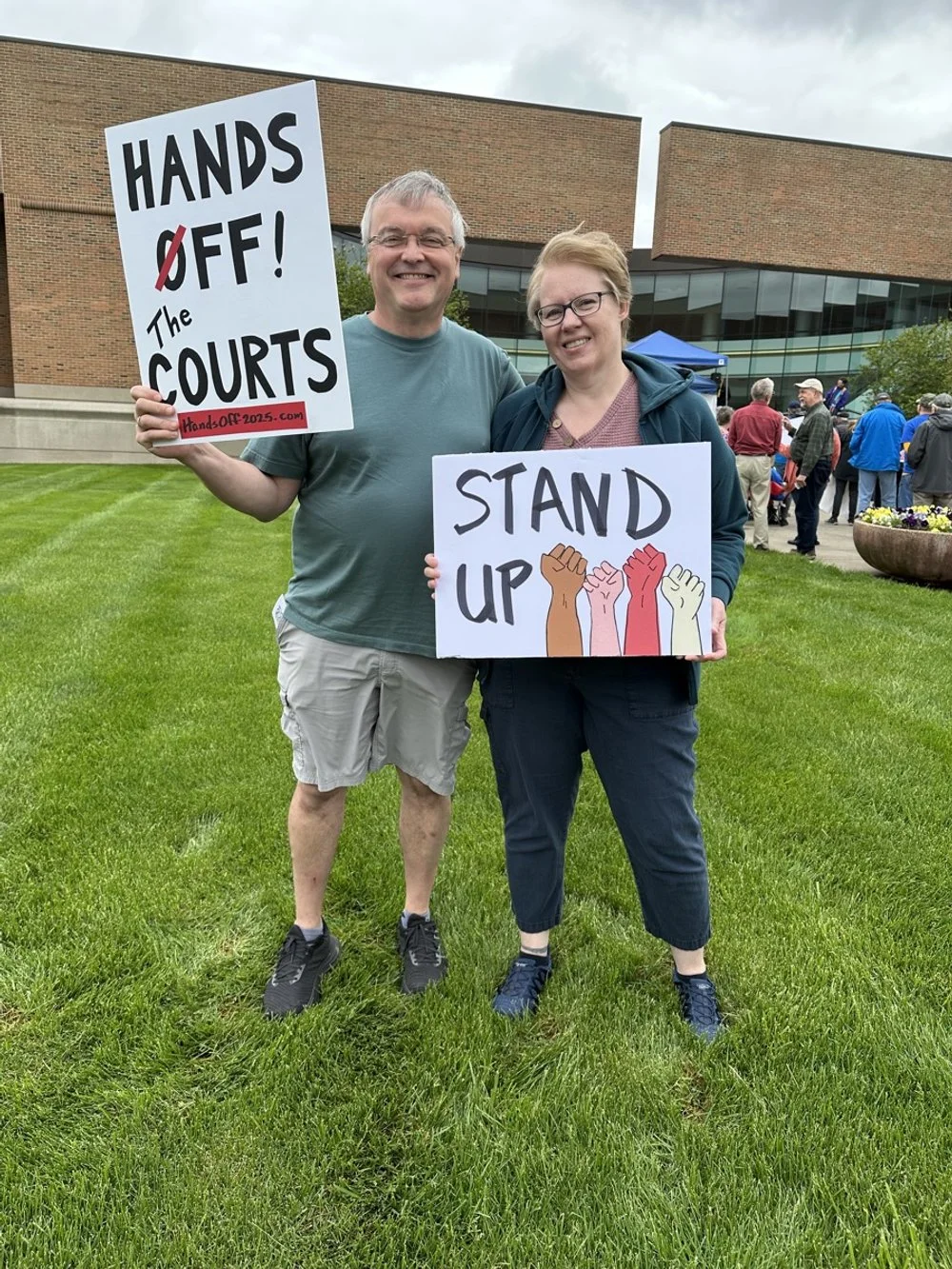 A man and a woman standing on grass in front of a modern building, holding protest signs that say 'HANDS OFF! The COURTS' and 'STAND UP' with diverse-colored raised fists.