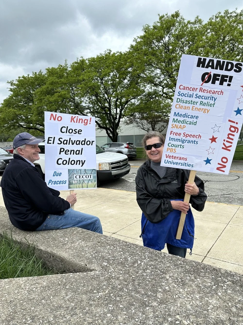Two people protesting outside; one is seated holding a sign against closing El Salvador Penal Colony, the other is standing holding a large sign with a list of issues and demands with slogans like "HANDS OFF!" and "No King!"