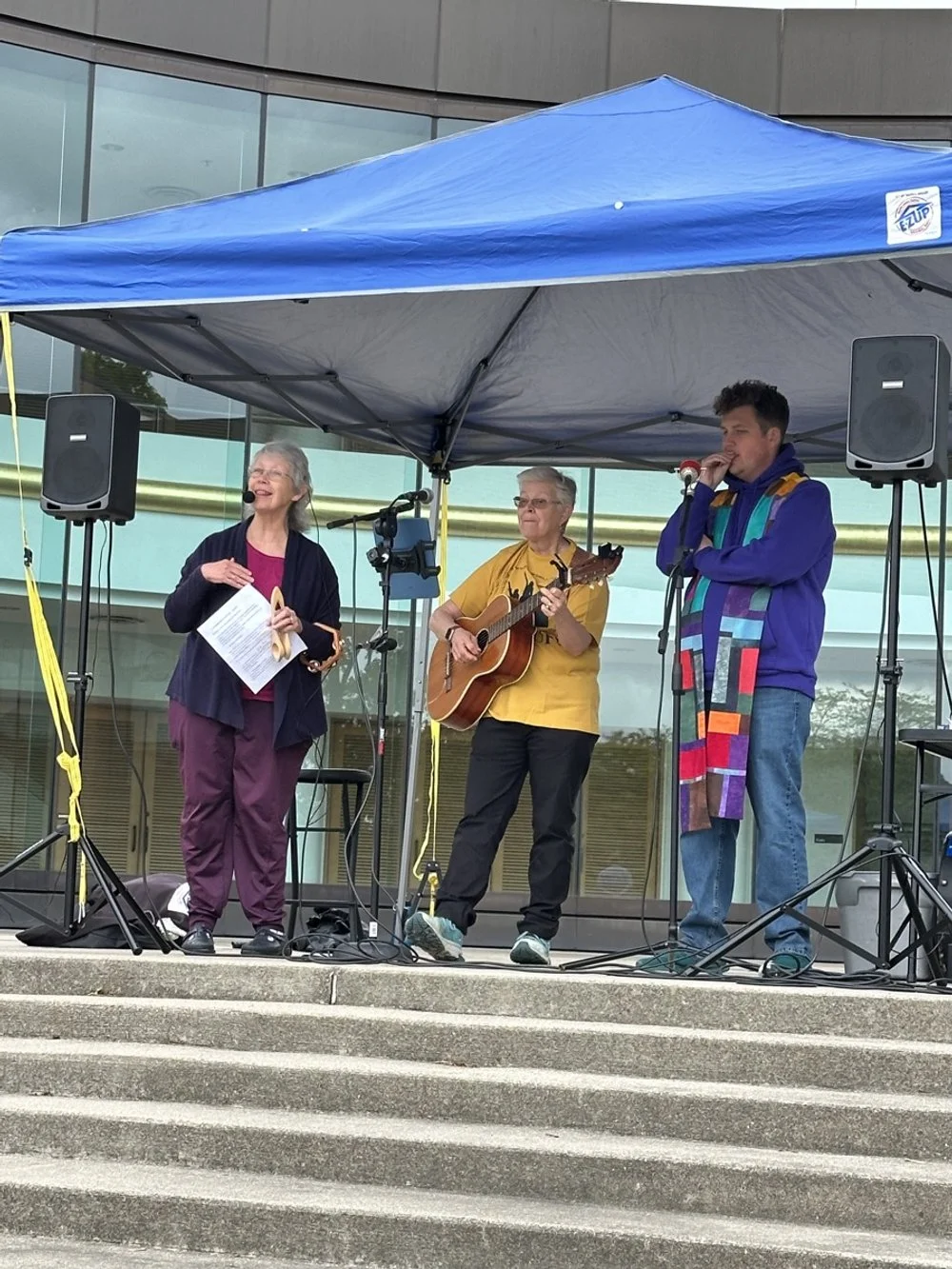 Three people performing on an outdoor stage under a blue canopy, with two speakers and musical equipment, in front of a modern glass building.