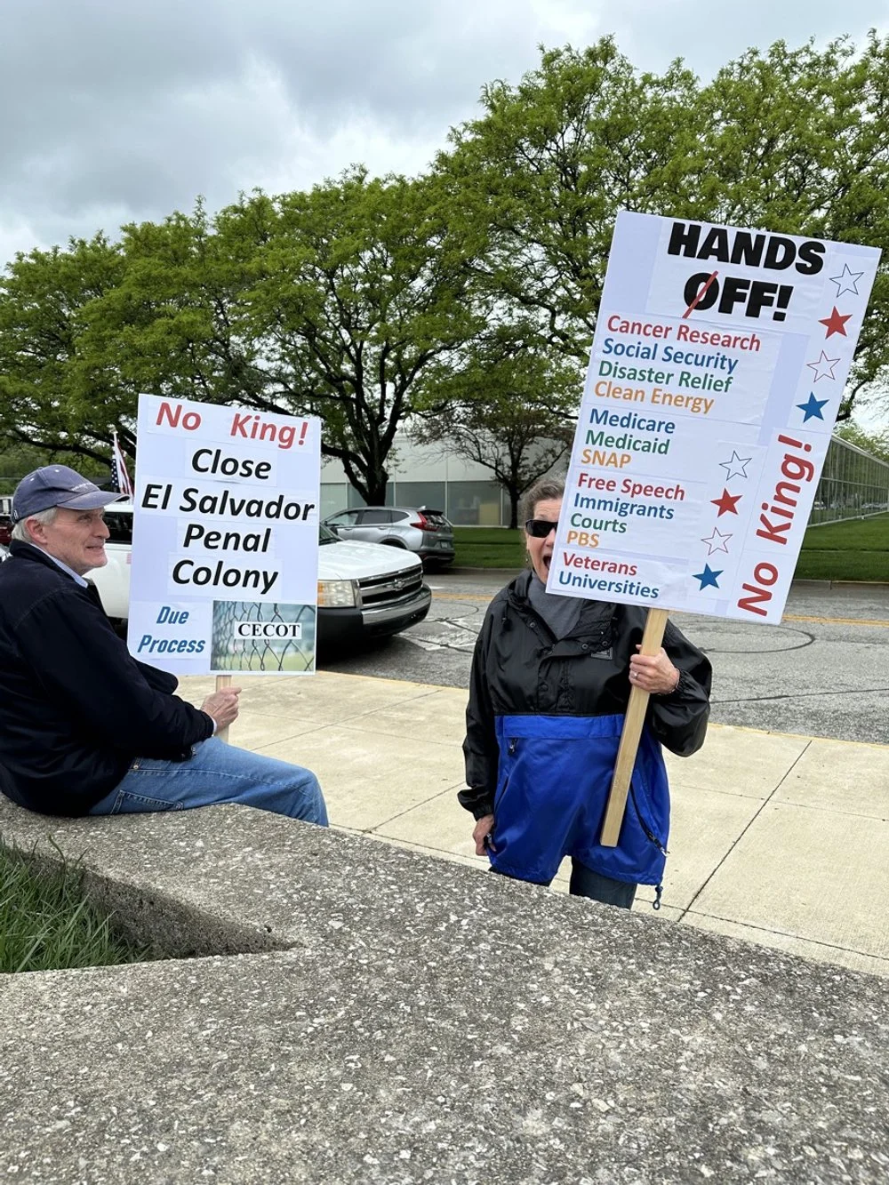 Two people holding protest signs outdoors on a cloudy day, with trees and parked cars in the background. One person is sitting on a concrete ledge, holding a sign protesting a close of El Salvador's penal colony, while another person stands, holding