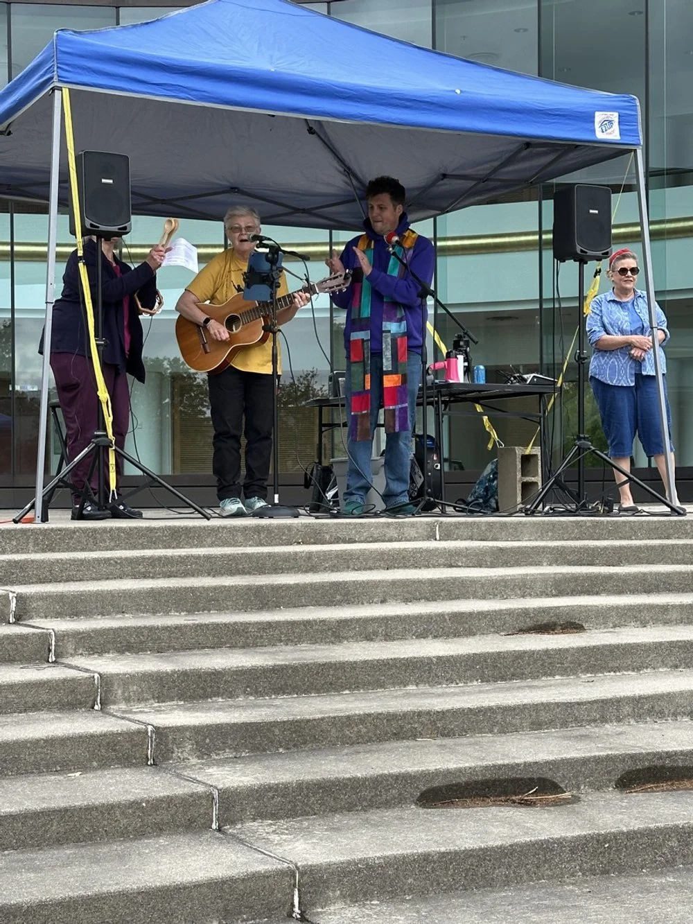 A group of four musicians performing on an outdoor stage under a blue canopy, with one playing guitar, others singing and playing instruments, in front of a modern building.