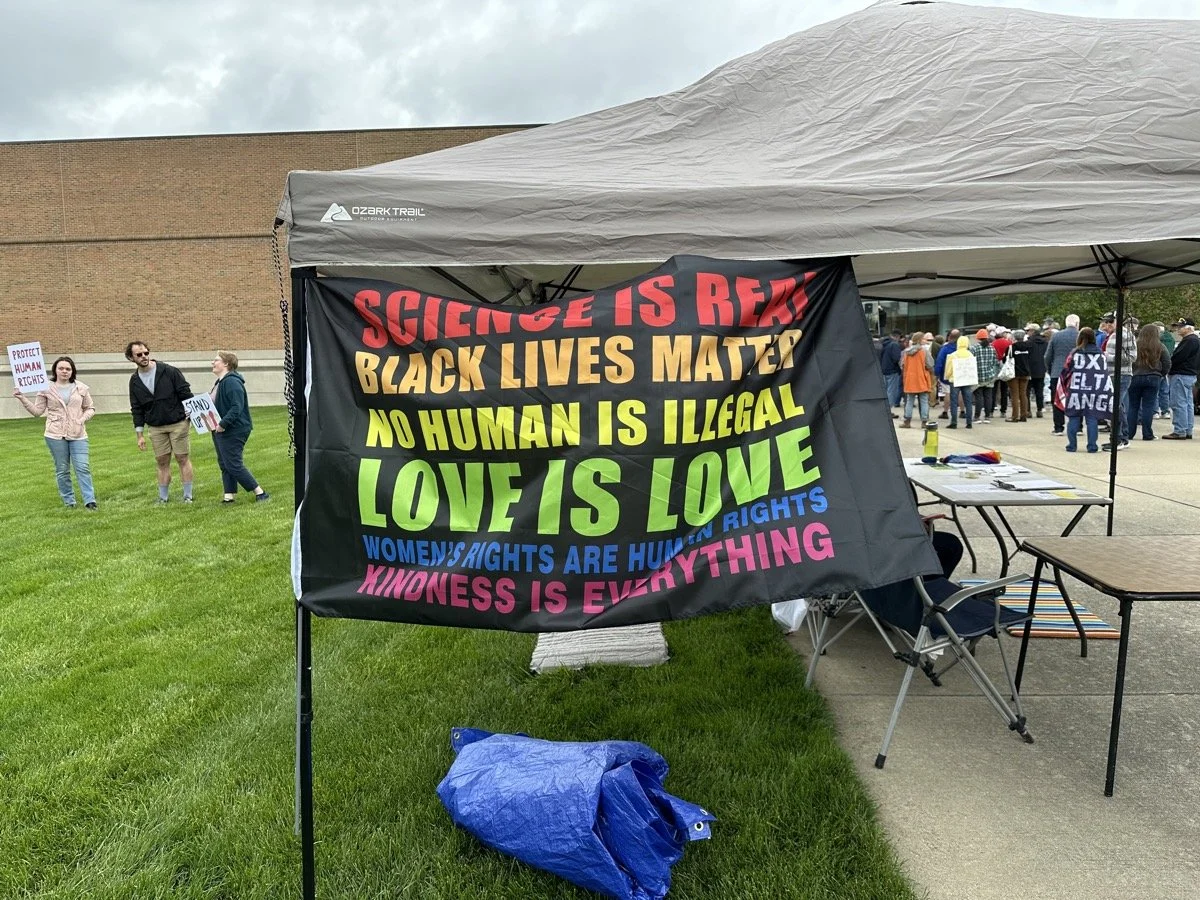 A protest gathering with a large banner promoting social justice messages, people holding signs, and a tent set up outdoors on a grassy area near a building.