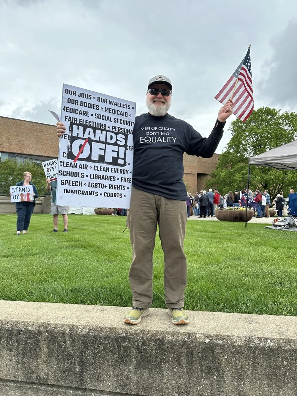 A man with a gray beard and sunglasses, smiling, holding a sign that says "HANDS OFF!" with a list of political issues, and an American flag, participating in a protest or rally outdoors with other people, some holding signs and a tent in the backgro
