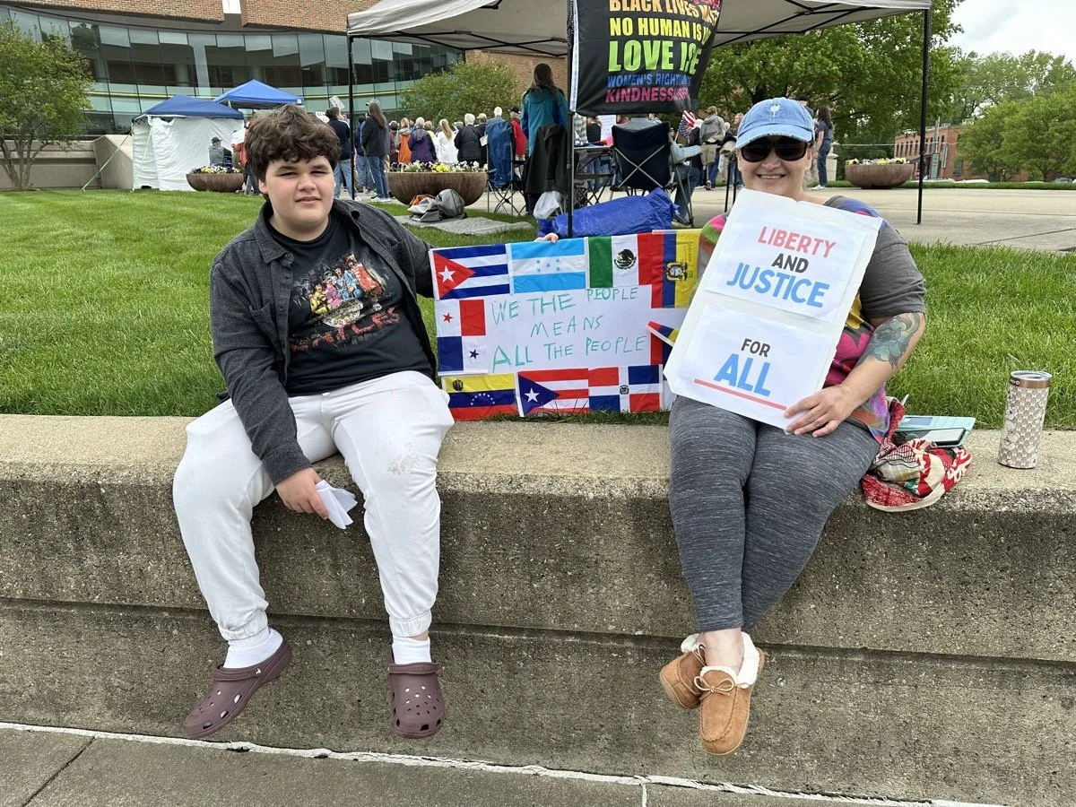 Two people sitting on a concrete ledge at an outdoor event, holding signs and banners advocating for justice and equality, with a crowd in the background.