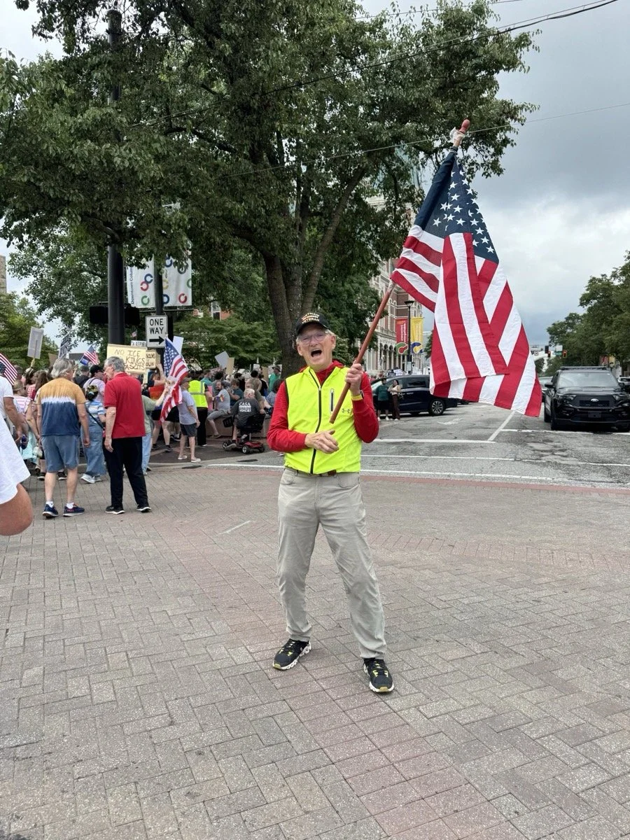 A man smiling and enthusiastically waving an American flag at a large outdoor gathering or protest. He is wearing a yellow vest over a red shirt and a black cap. In the background, there are many people holding smaller flags and signs, with some tree