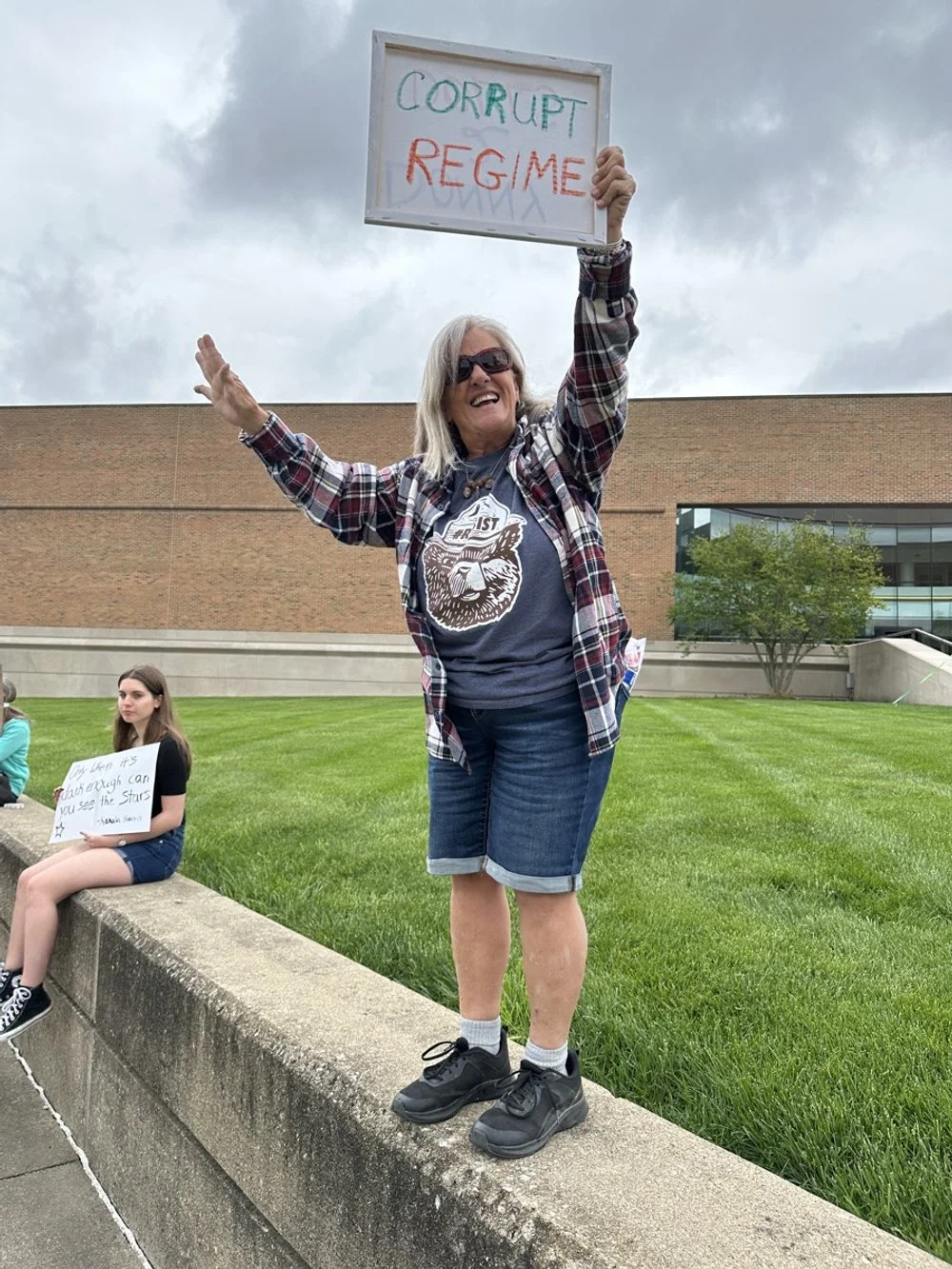 Woman smiling, holding a sign that reads "CORRUPT REGIME" during a protest or demonstration outside. Other people are seated nearby, one holding a sign.
