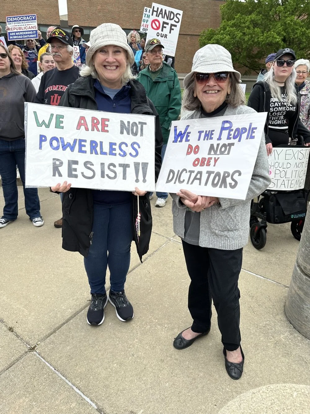 Two women holding signs at a protest or rally, smiling at the camera. The woman on the left holds a sign that reads, "WE ARE NOT POWERLESS RESIST!" The woman on the right holds a sign that says, "WE THE PEOPLE DO NOT OBEY DICTATORS." Other protesters