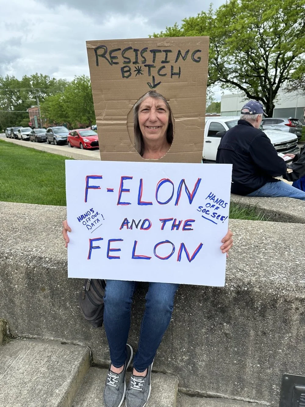 A woman holding a protest sign with a cardboard cutout face hole and a sign with the message "FELON AND THE FELON" and handwritten notes saying "HANDS OFF OUR DATA!" and "HANDS OFF OUR SOCIAL SECURITY!". She is standing outdoors on a sidewalk with tr