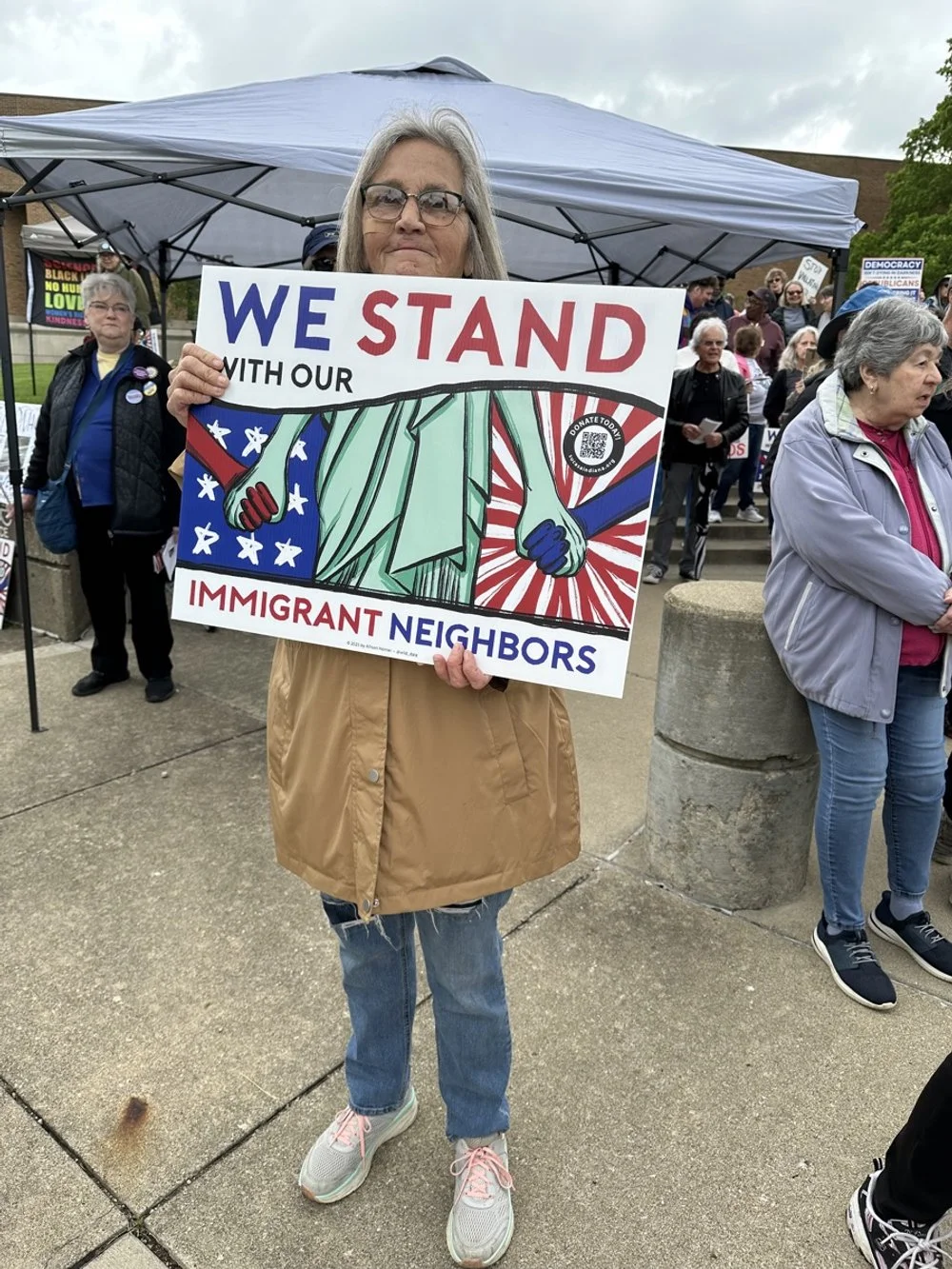 A woman holding a sign that reads "We Stand with Our Immigrant Neighbors" during a protest or rally. There are other people and signs in the background, and a blue tent overhead.