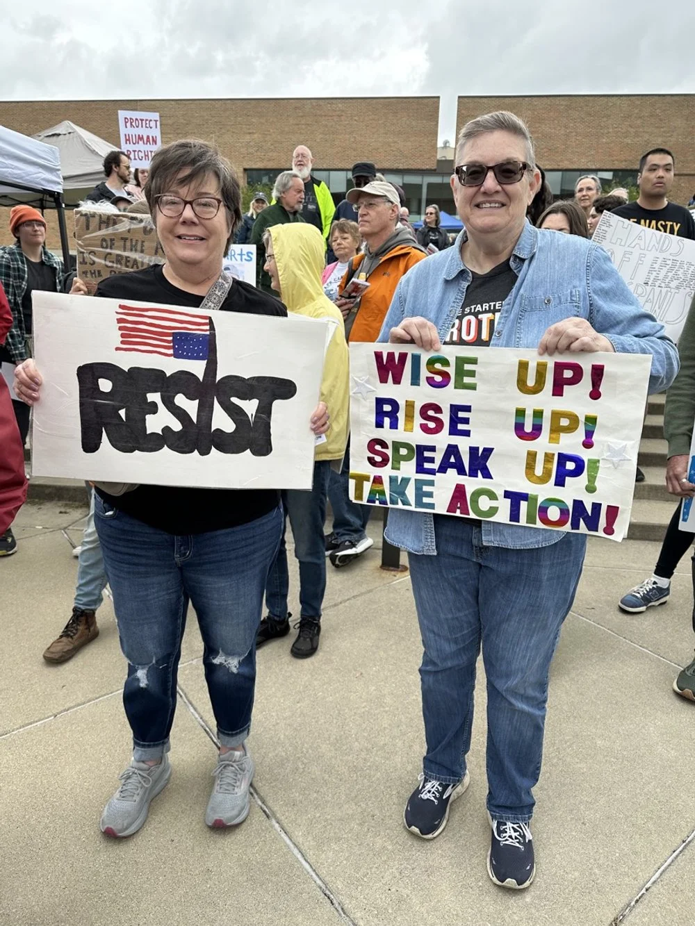 Two women protesting at a rally holding colorful signboards. One sign reads 'RESIST' with an American flag painted in the shape of a fist, and the other reads 'WISE UP! RISE UP! SPEAK UP! TAKE ACTION!' surrounded by several other protesters holding s