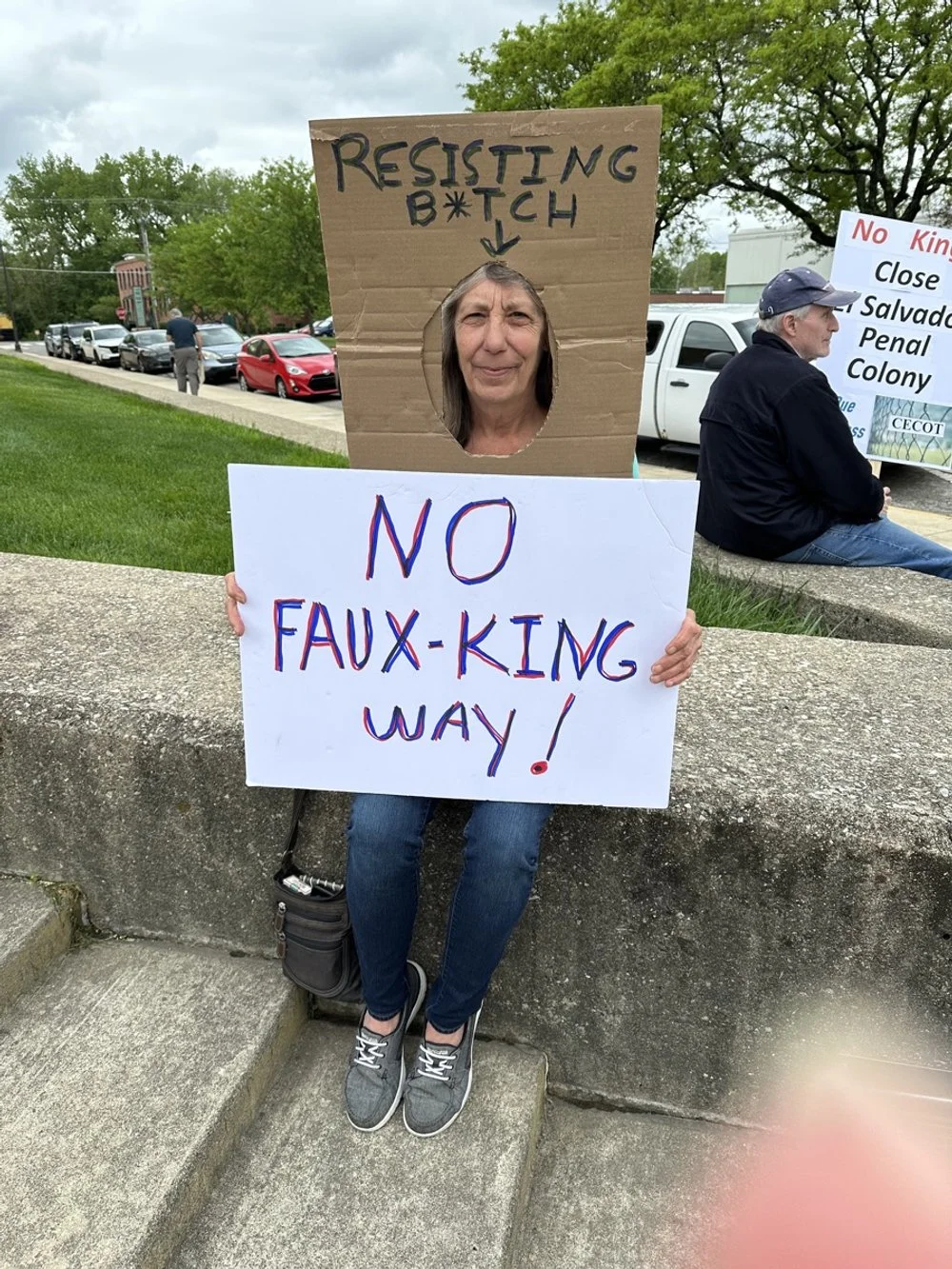 A woman sitting on a concrete ledge holding a white protest sign that reads "NO FAUX-KING WAY!" with a cardboard cutout mask around her face, which has a sign above it reading "RESISTING B*TCH." In the background, there are people sitting and cars pa