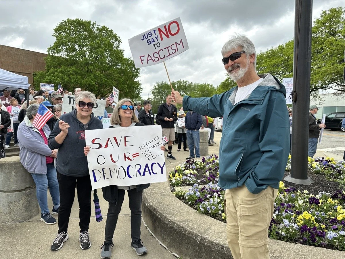 Group of people at a protest or rally holding signs that say "Save our democracy" and "Just say no to fascism." The signs advocate for democracy and oppose fascism. The crowd includes individuals of various ages, some wearing sunglasses and holding A
