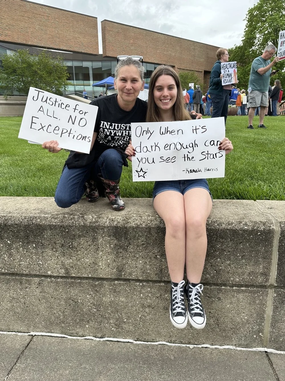 A young woman sitting on a concrete ledge holding a sign that reads, 'Only when it's dark enough can you see the stars – Kamala Harris.' Next to her, a woman crouching holding a sign that says, 'Justice for all. No exceptions.' They are at a protest