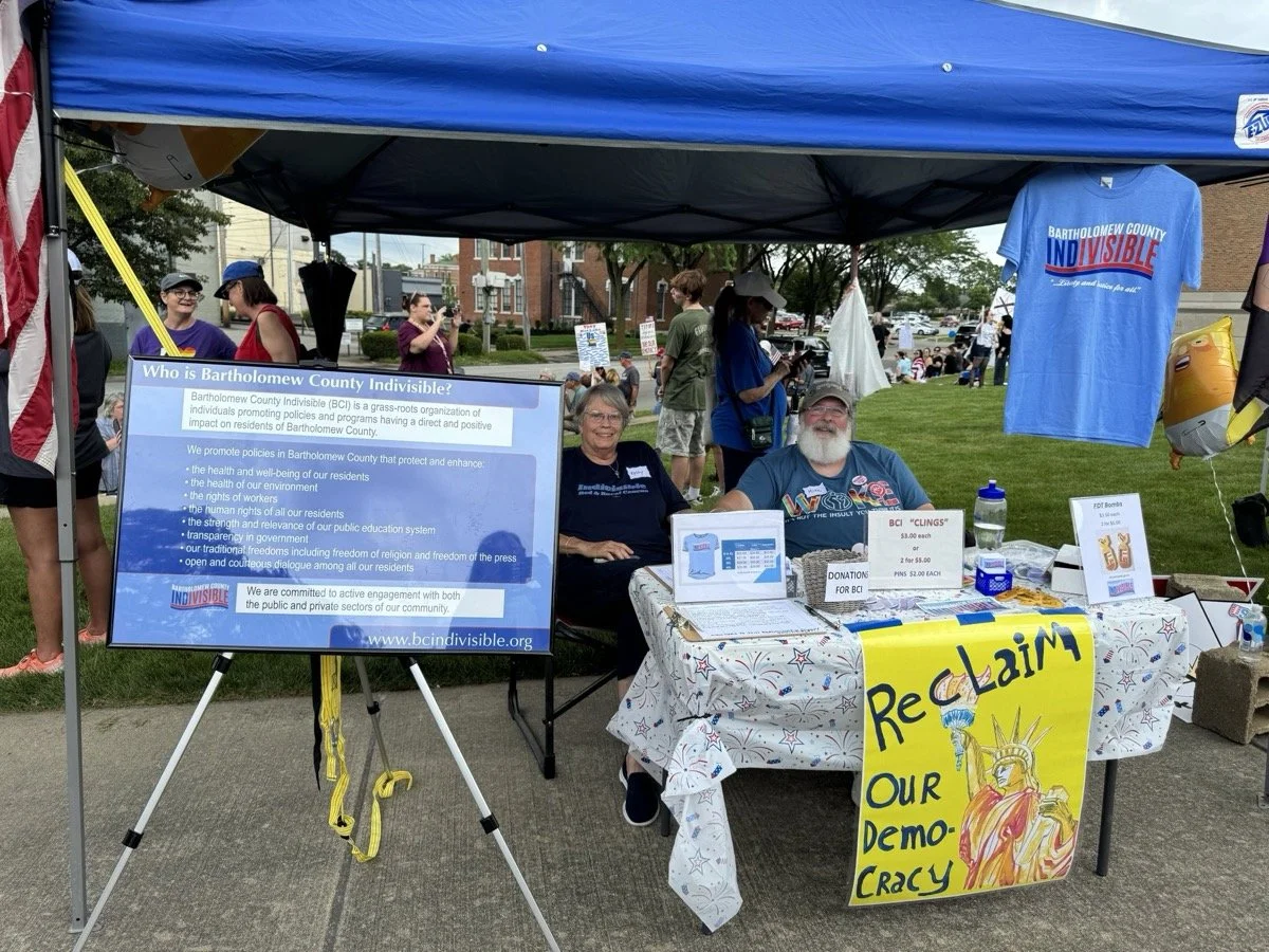 Booth at an outdoor event with a large informational sign about Bartholomew County Indivisible, two smiling people sitting behind the table, and a yellow poster in front that reads 'Reclaim Our Democracy,' featuring a drawing of the Statue of Liberty