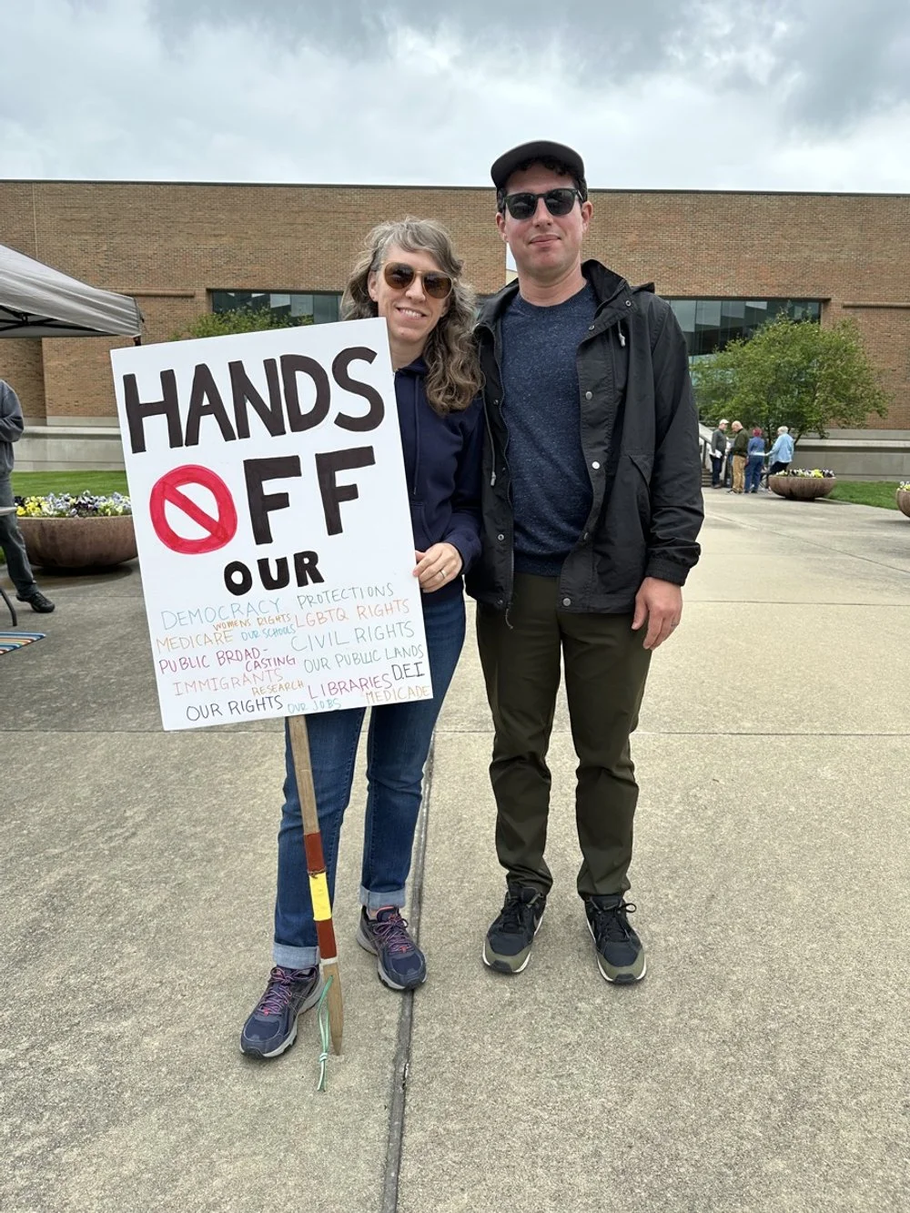 Two people standing outdoors, holding a sign protesting against certain political issues. The woman on the left has gray hair and sunglasses, and the man on the right has dark hair, sunglasses, and a black jacket. There are other people and a buildin
