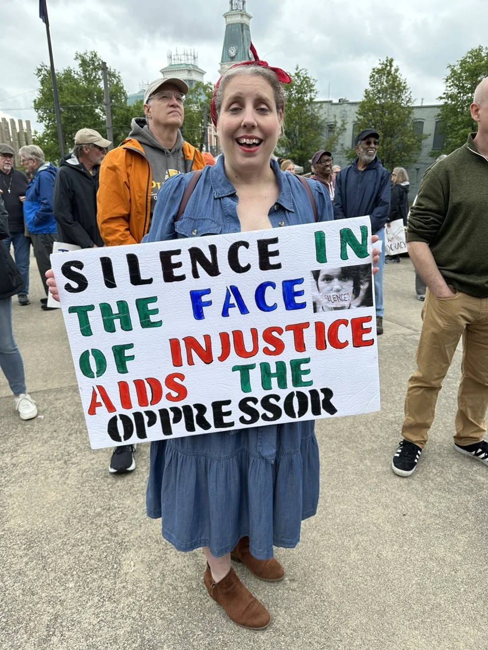 A woman holding a sign at a protest or rally with other people in the background. The sign reads, 'Silence in the face of injustice AIDS the oppressor' with the words 'injustice' and 'oppressor' in red and others in black, green, blue, and red.