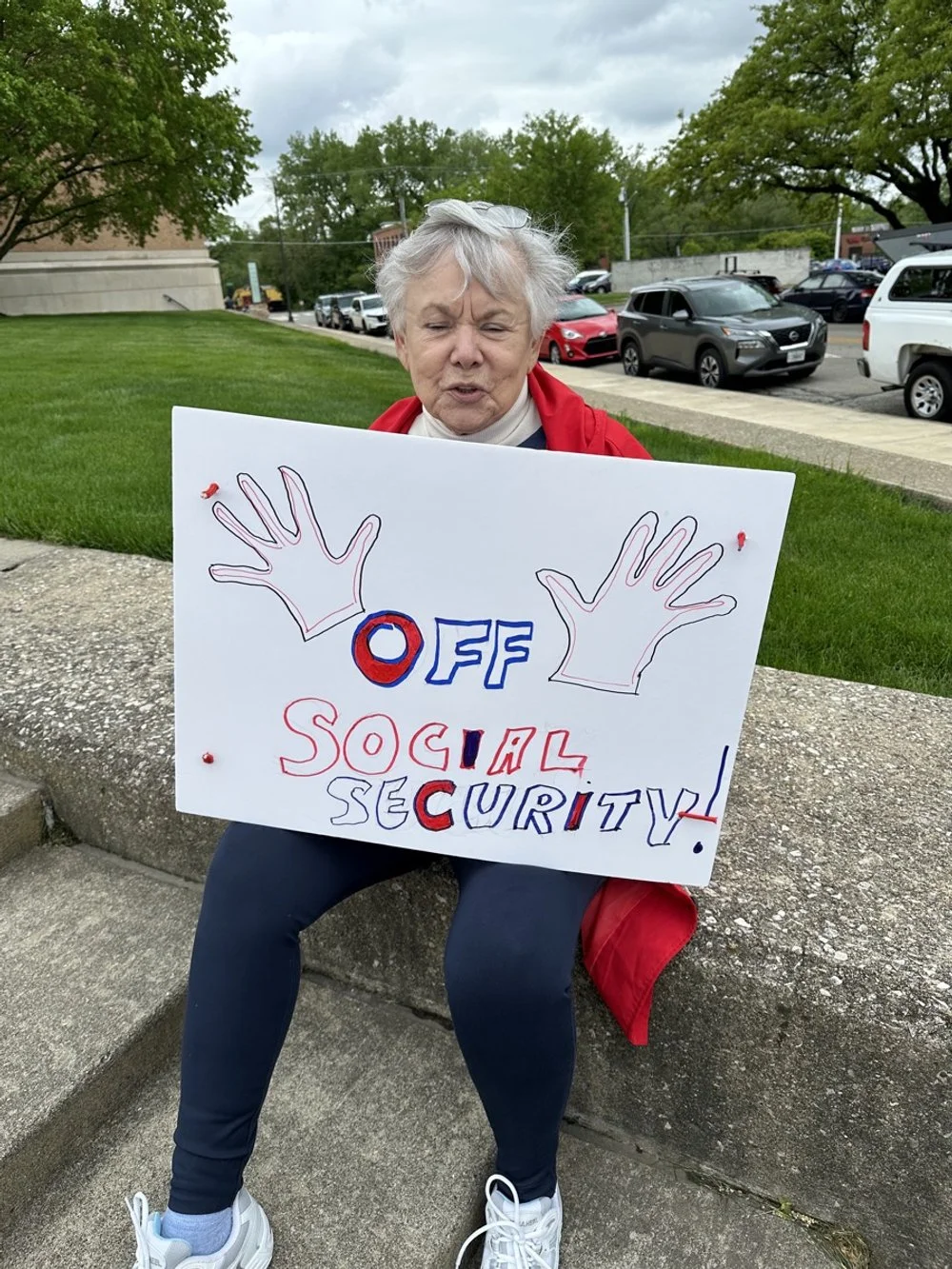 An elderly woman sitting on a stone bench outdoors holding a protest sign that reads "OFF SOCIAL SECURITY" with cut out hand illustrations and red, white, and blue lettering.