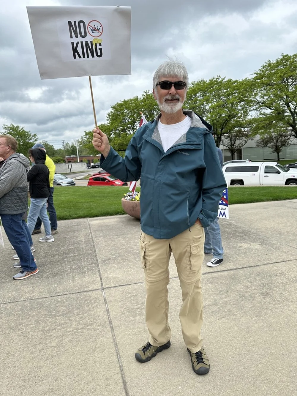 A man with gray hair and beard, wearing sunglasses, a teal jacket, and beige pants, holds a sign that says "NO KING" with an image of a crown crossed out. He is outdoors on a cloudy day, standing on a sidewalk with people in line or gathering behind