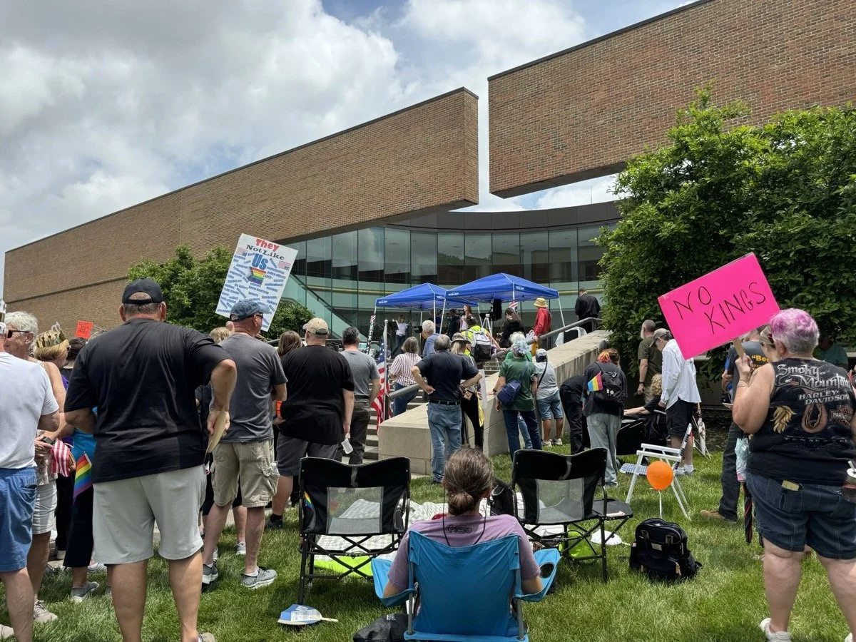 A crowd gathered outside a modern building at a protest or rally, with signs including 'NO KINGS' and 'They Not Like Us', some people seated and others standing, with tents and a stage in the background.