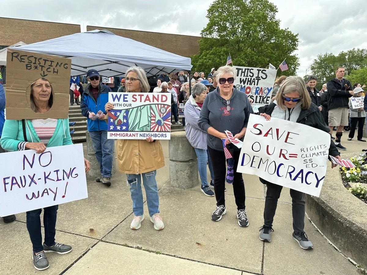 Group of people holding protest signs at an outdoor rally, some signs read 'No Faux-King Way!', 'We Stand with Our Immigrant Neighbors', and 'Save Our Democracy'.