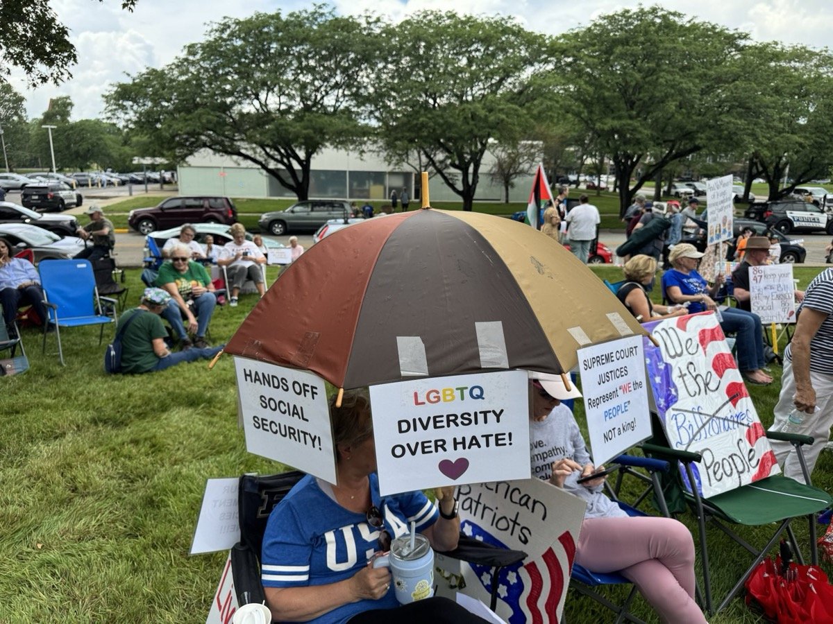 People protesting at a rally holding signs with messages advocating for LGBTQ rights and diversity, sitting outdoors on chairs with trees and cars in the background.