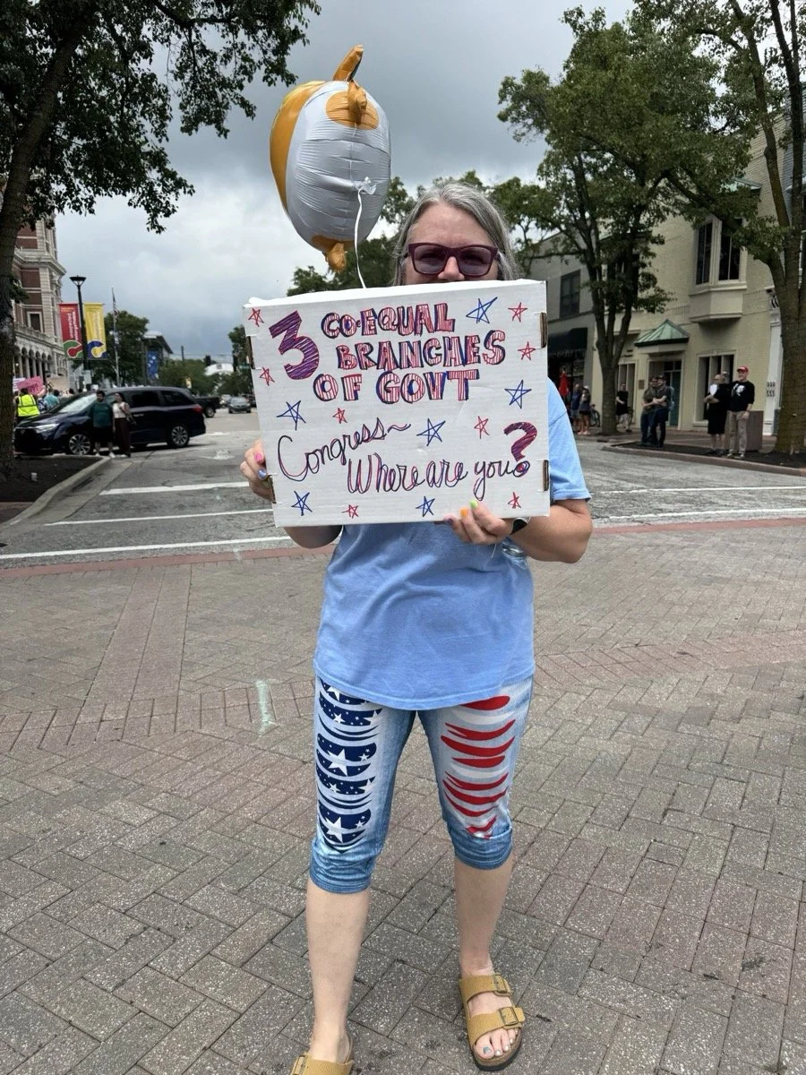 Woman wearing sunglasses and patriotic leggings holding a sign that reads '3 Coequal Branches of Government Congress Where are you?' standing on a city sidewalk. A unicorn-shaped balloon is above her. There are people and buildings in the background.