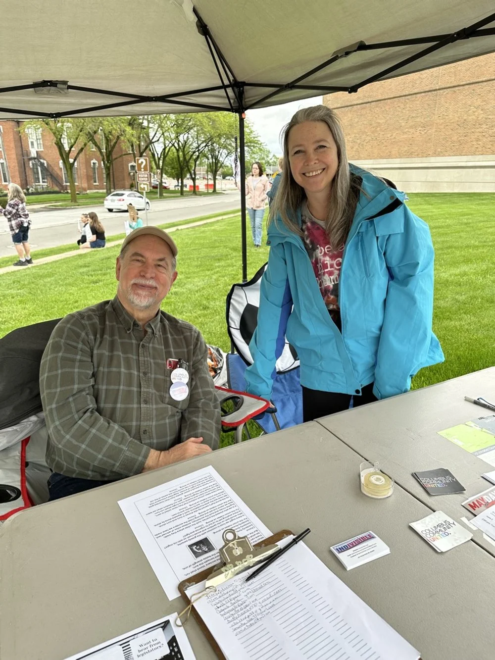 A woman in a blue jacket standing beside a seated man at an outdoor event table under a canopy, with a grassy area, trees, and a brick building in the background.