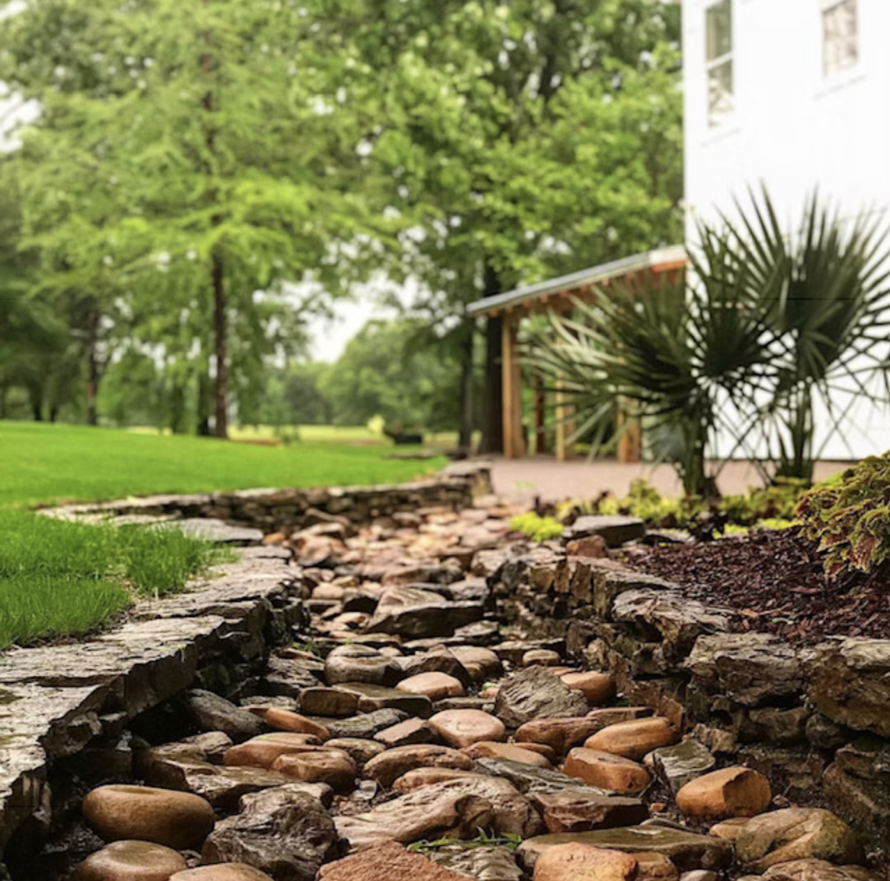 A landscaped backyard with a stone-lined dry creek bed, green grass, trees, and a white house with a wooden porch.