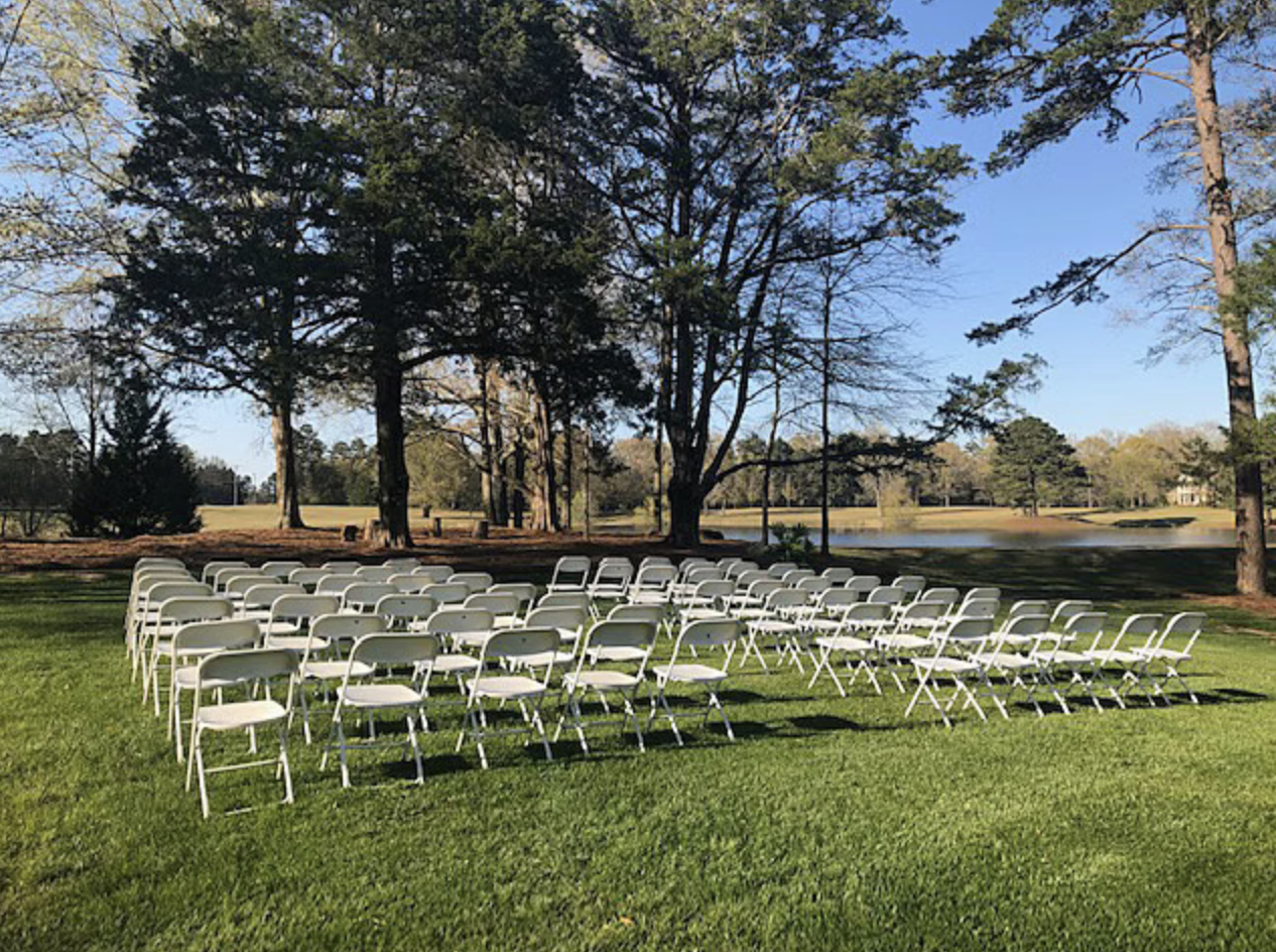 White chairs arranged in rows on green grass in an outdoor park near a lake, surrounded by trees under a clear blue sky.