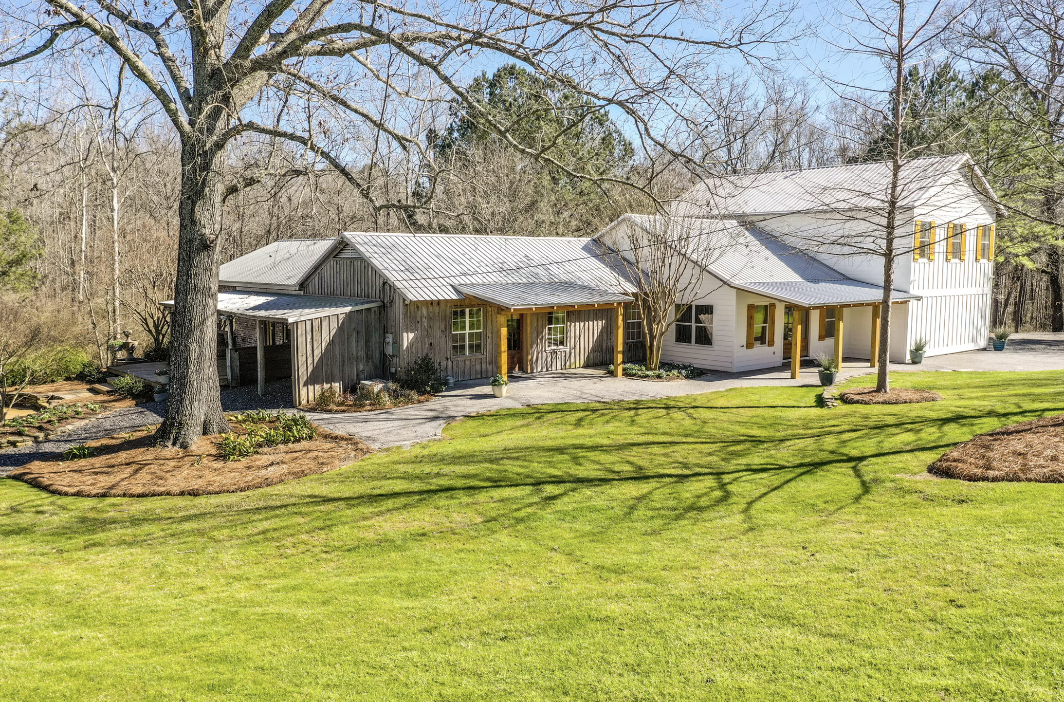 A house with a gray metal roof, a wooden and white exterior, surrounded by a well-maintained lawn, trees, and garden beds in a rural setting.