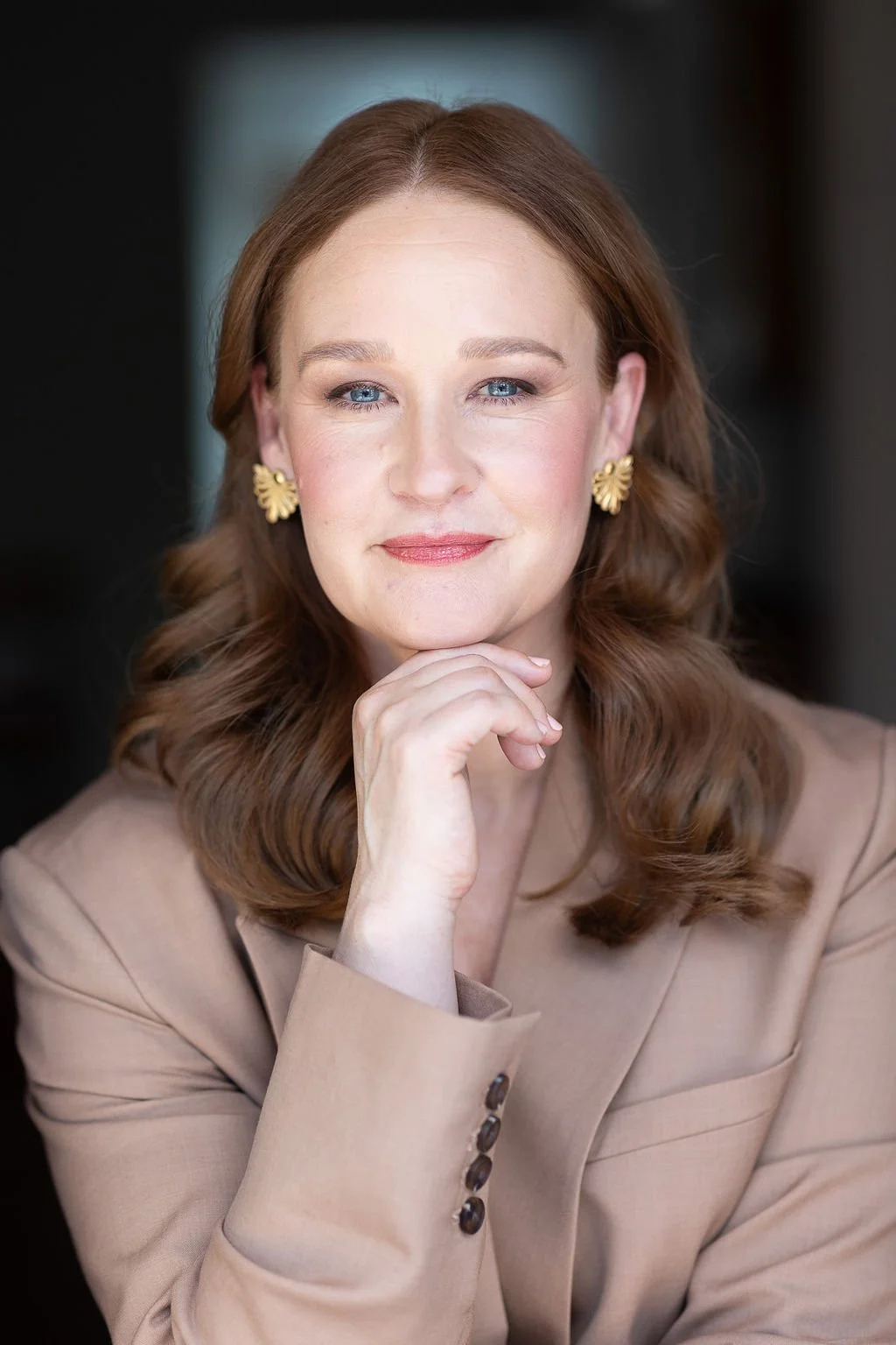 A woman with red hair and blue eyes, wearing gold earrings and a beige blazer, resting her chin on her hand and smiling slightly.