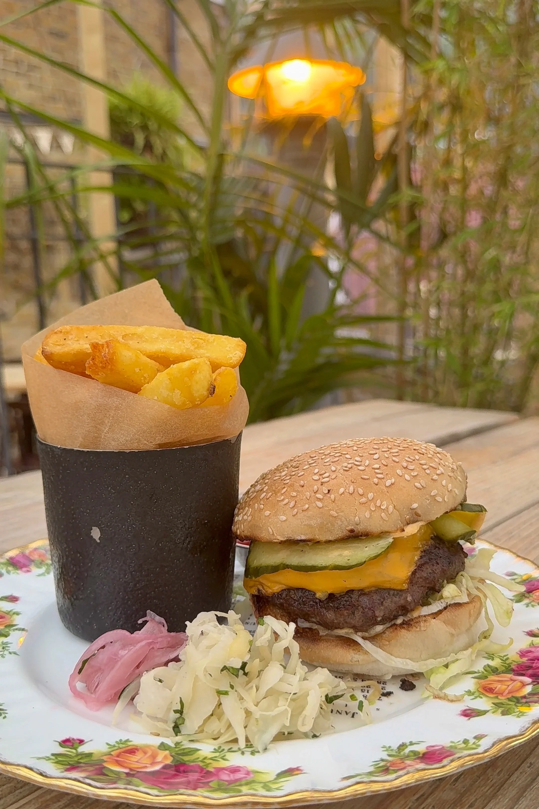 A plate with a hamburger, French fries in a black container, and some coleslaw on a wooden table outdoors