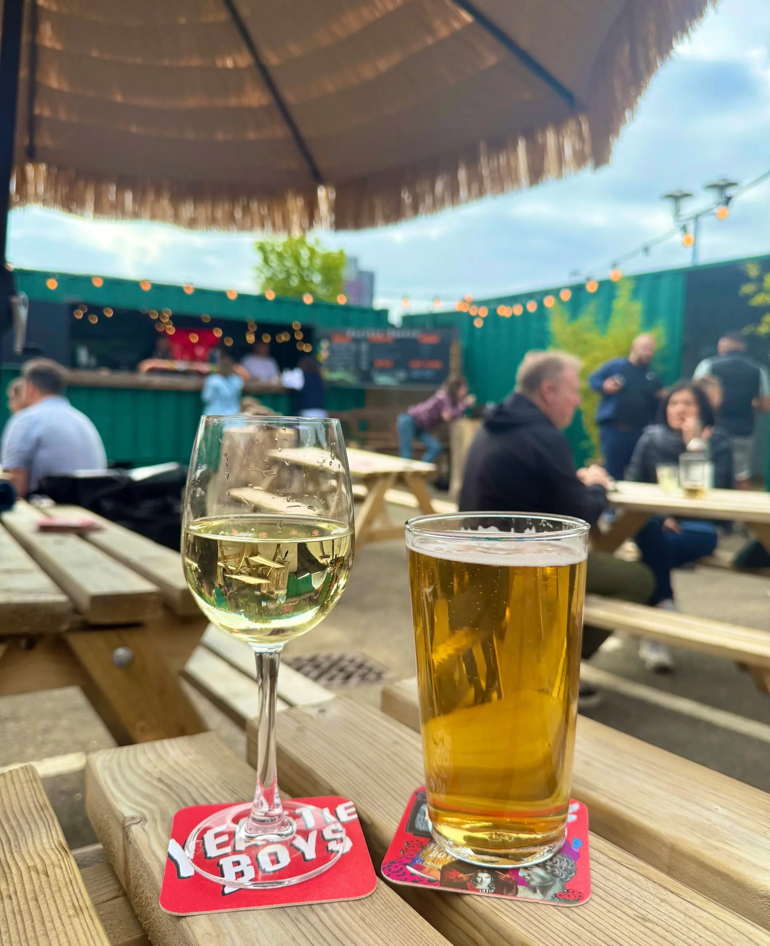A glass of white wine and a glass of beer on a wooden picnic table at an outdoor bar with people socializing in the background.
